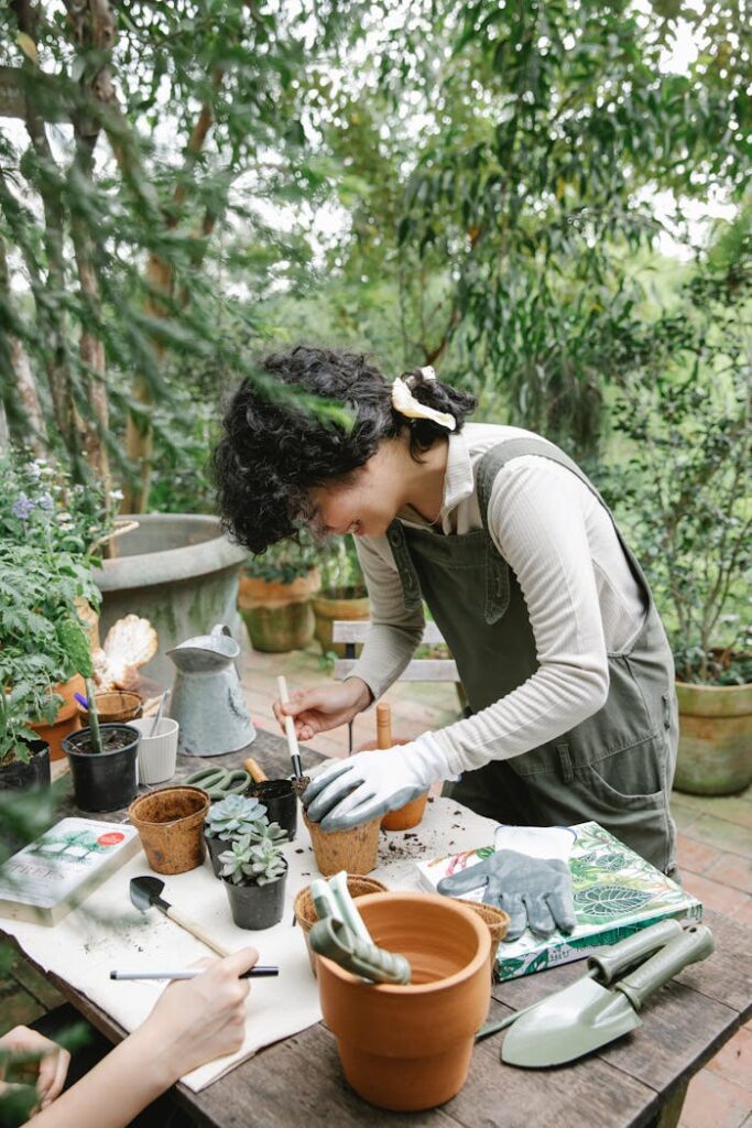 Side view of concentrated ethnic female gardener standing at wooden table with gardening tools while planting flower in pots during work in orangery